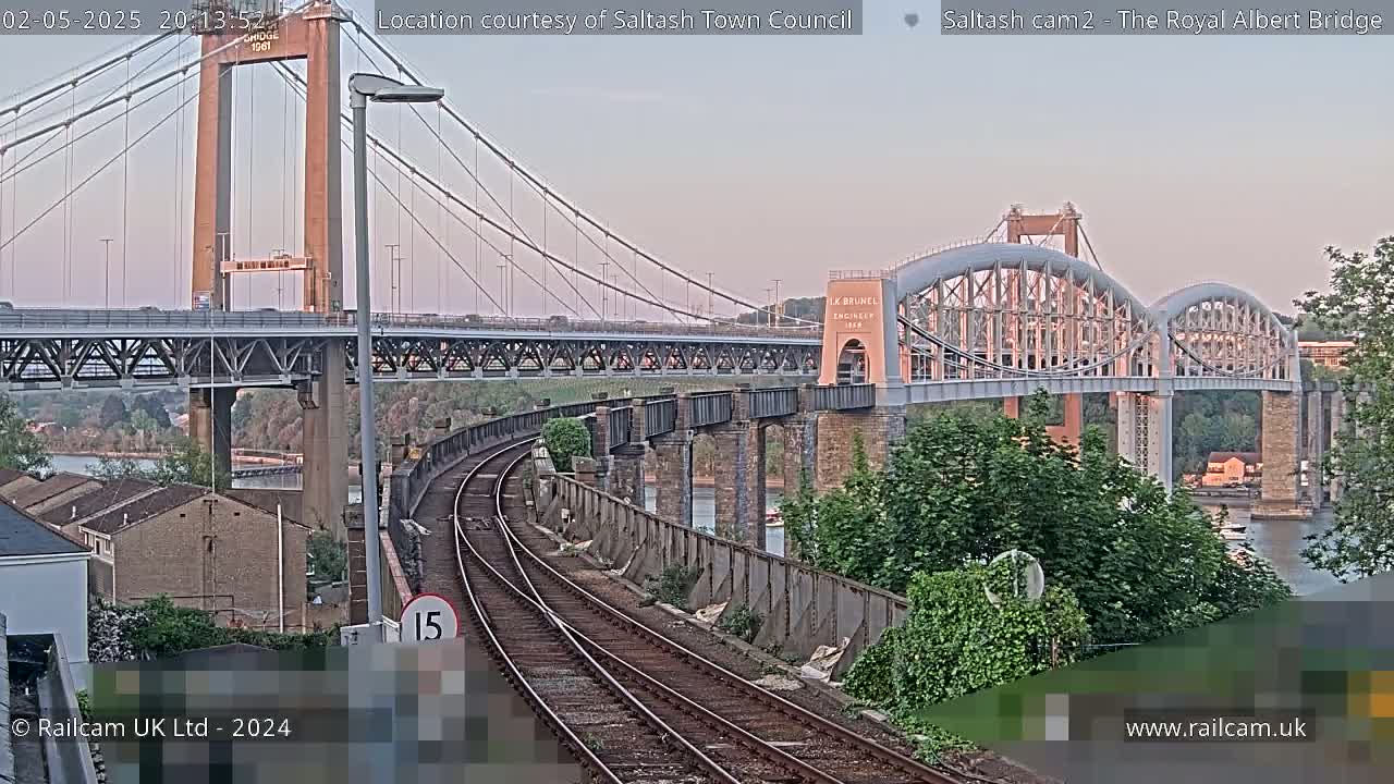A view of railroad tracks leading to a large suspension bridge and an arched bridge over water, under a twilight sky.