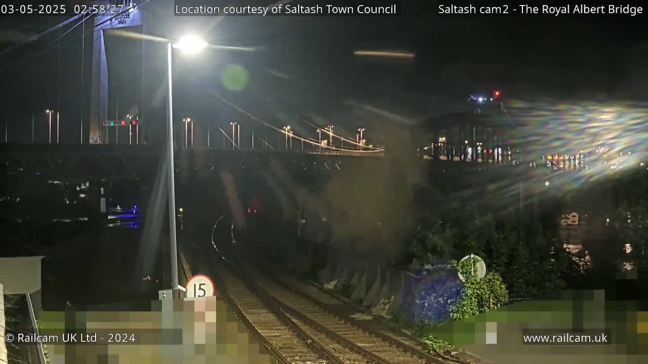 A nighttime view of a suspension bridge and railway tracks, with a train emerging from a tunnel.