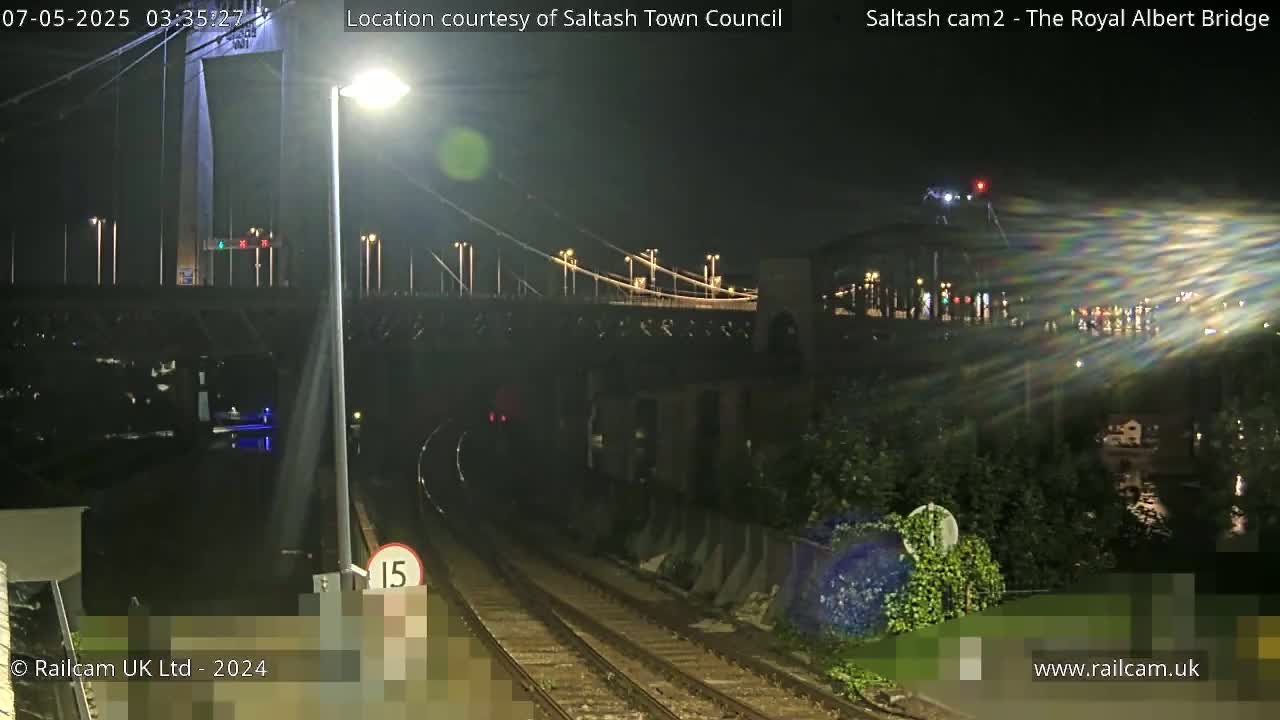 A nighttime view of a railway line leading towards a suspension bridge and another bridge in the distance, under a clear sky.
