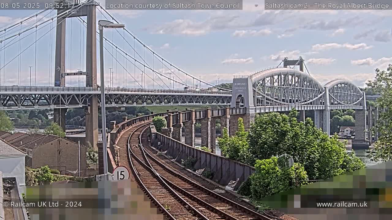 A train track curves alongside a river, under a suspension bridge and a nearby arch bridge, on a partly sunny day.