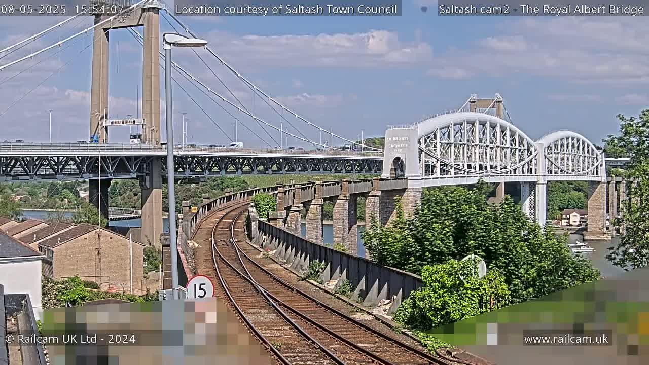 A railway track curves alongside a river, with a modern suspension bridge and an older arched bridge visible in the background under a partly sunny sky.