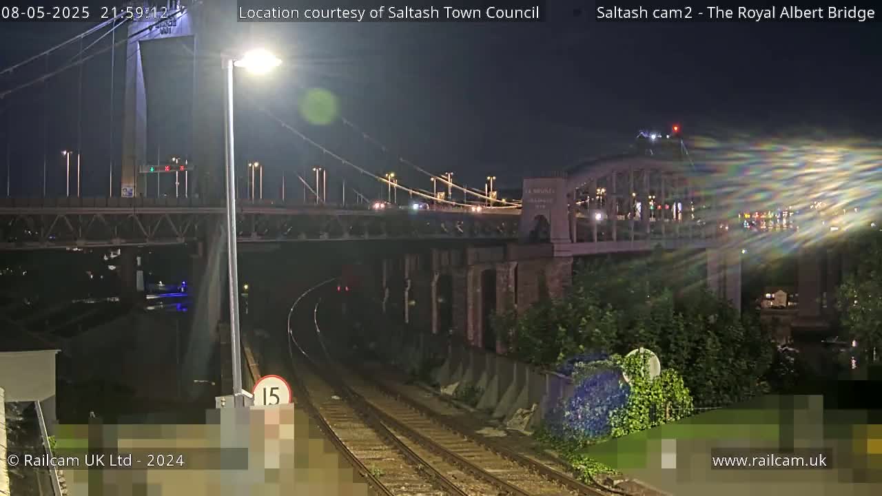 A nighttime view of a railway track leading toward a bridge over a body of water, with a second, larger bridge visible in the distance.
