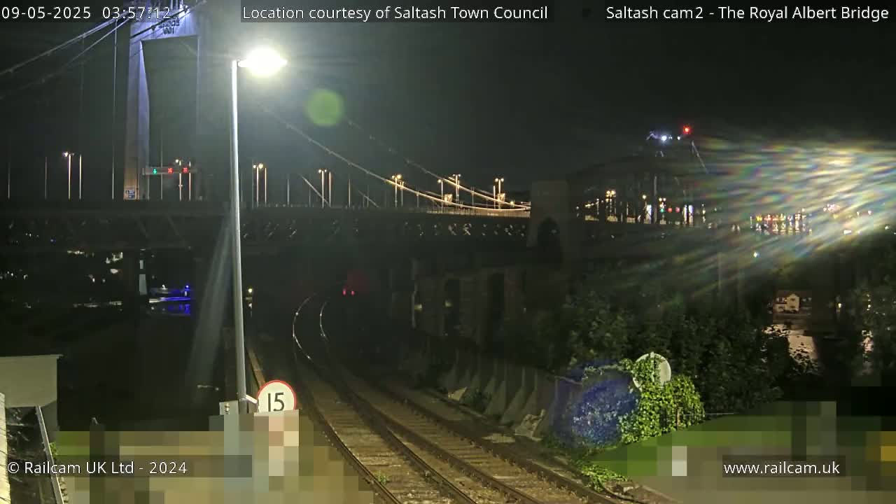 A nighttime view of a suspension bridge over railroad tracks, illuminated by streetlights.