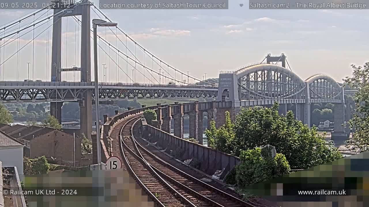 Two bridges, one a suspension bridge and the other an arch bridge, span a waterway under a mostly clear sky, with train tracks visible in the foreground.
