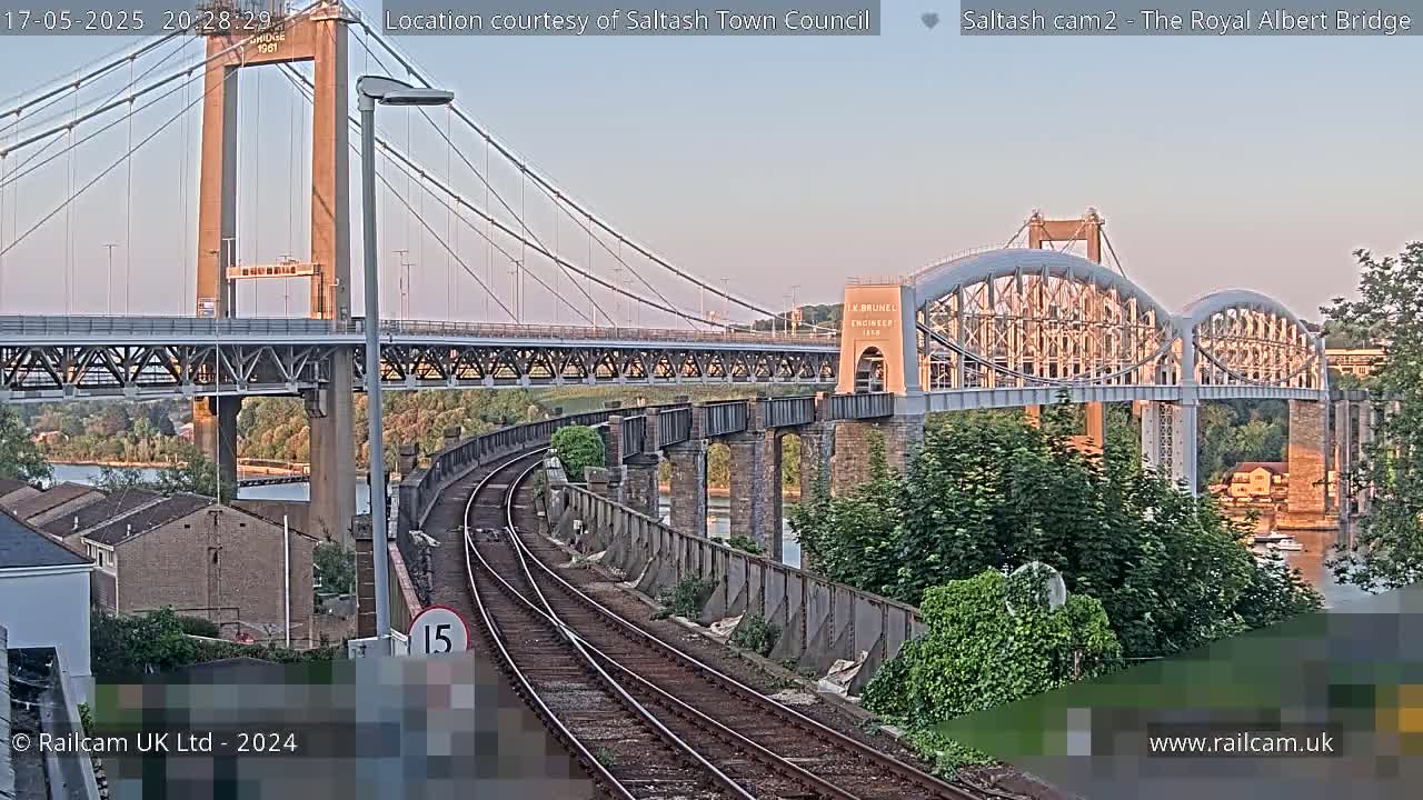 A railway track runs beside a river, under two bridges, one a suspension bridge and the other an arch bridge, at dusk.