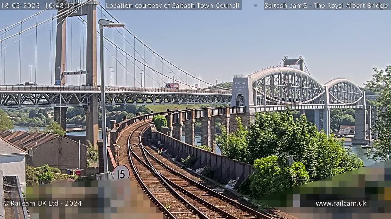 A sunny day reveals a railway track curving towards two bridges spanning a body of water, one a modern suspension bridge and the other an older arch bridge.