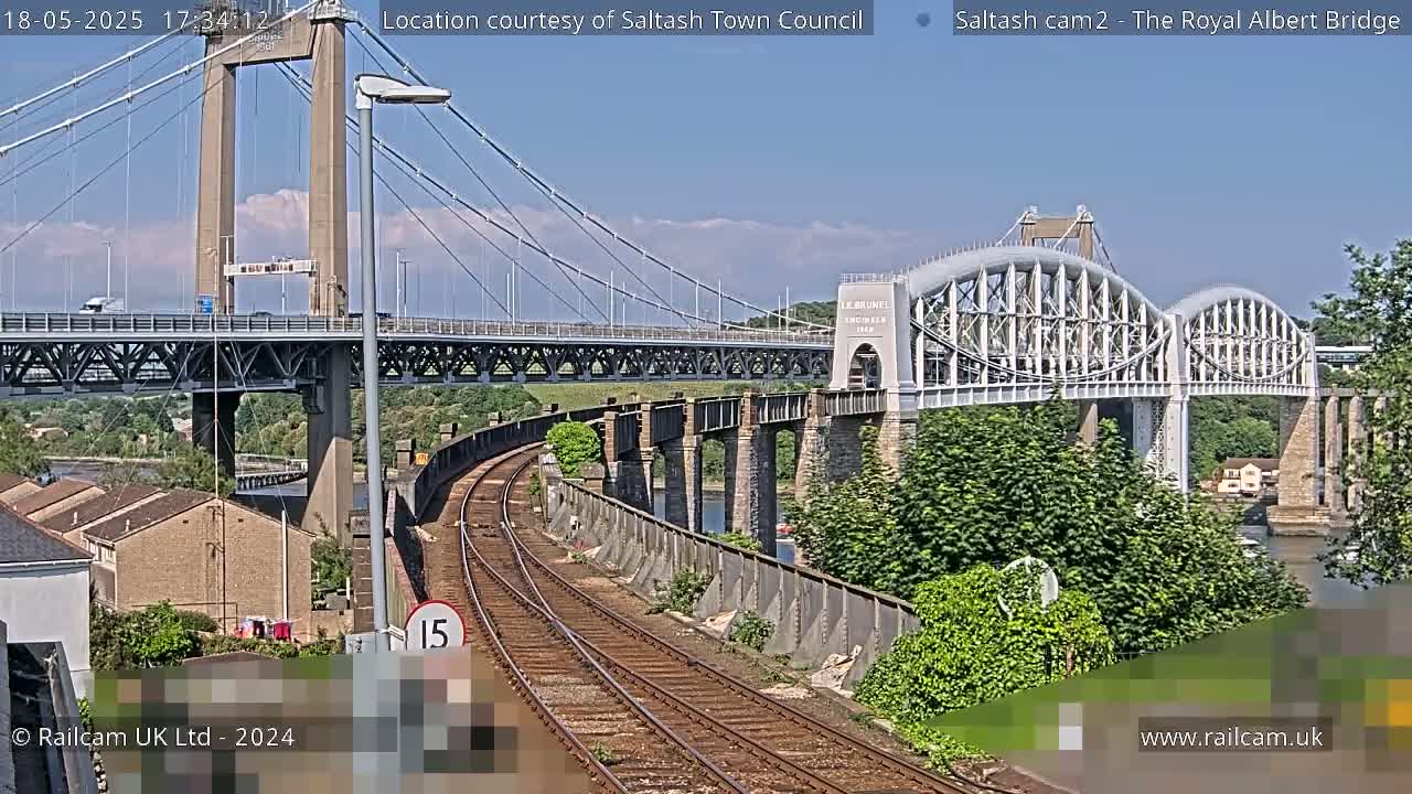 Under a sunny sky, a train track curves towards two bridges, one a suspension bridge and the other an arch bridge, with lush green vegetation lining the riverbank.