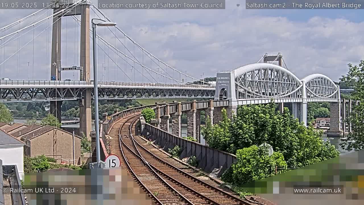 Train tracks curve towards two bridges, a modern suspension bridge and an older arched bridge, under a partly cloudy sky.