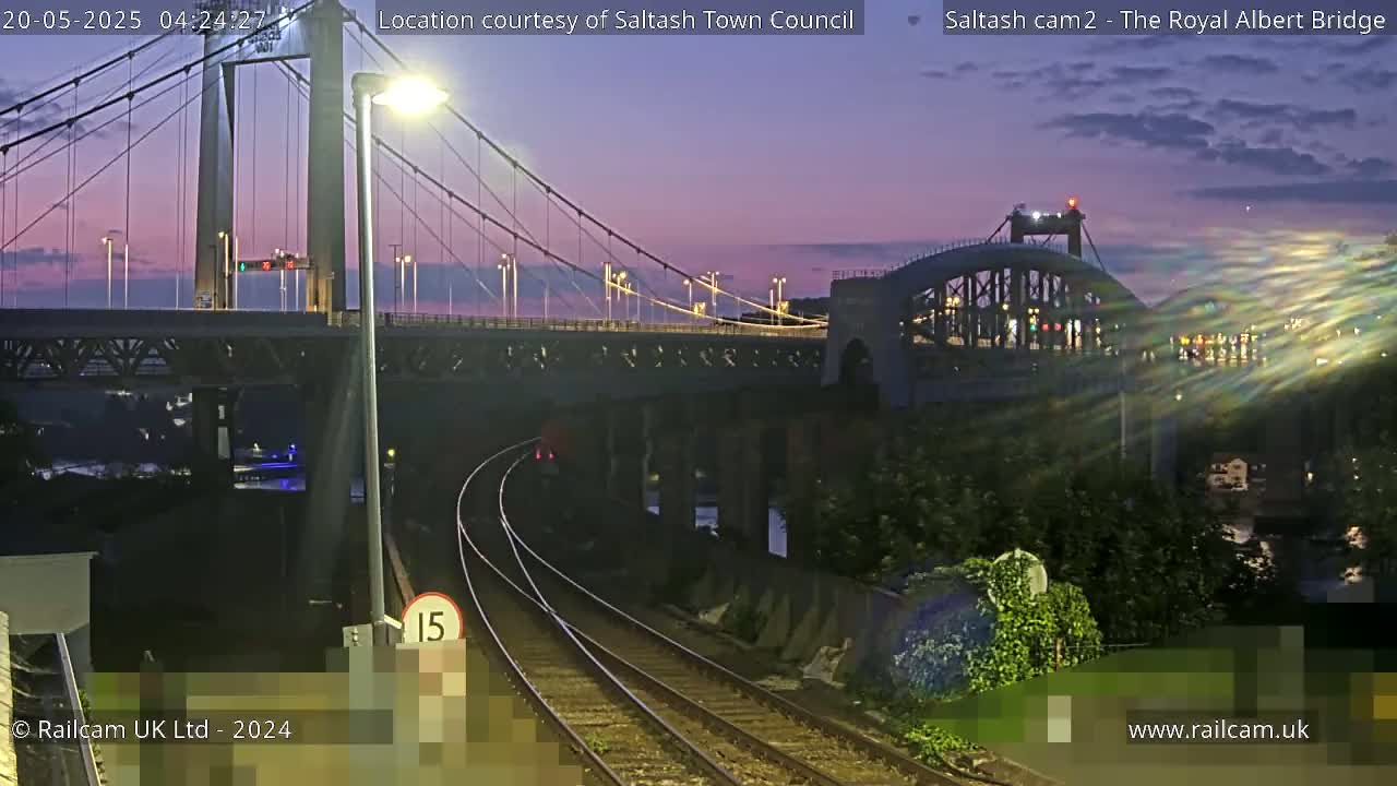 A twilight view of a railway line leading toward two bridges, one a suspension bridge and the other an arch bridge, under a dusky purple sky.