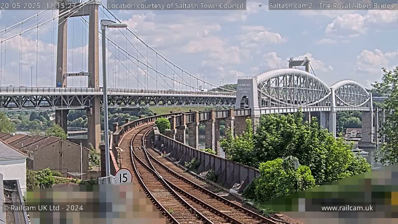 Under a partly cloudy sky, a railway line curves alongside a suspension bridge and an older arch bridge, with lush green trees visible in the background.