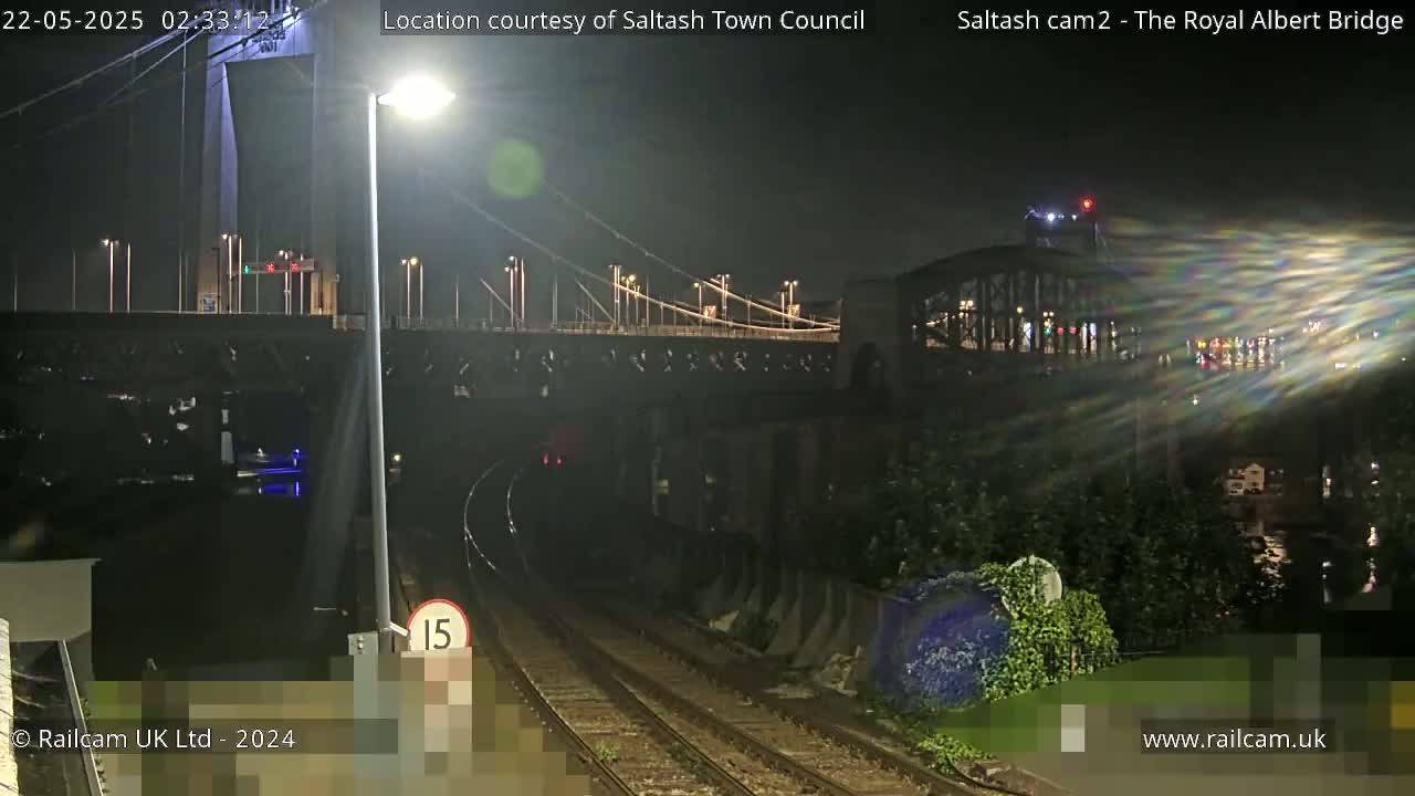 A nighttime view of a train track leading under a suspension bridge, illuminated by streetlights.