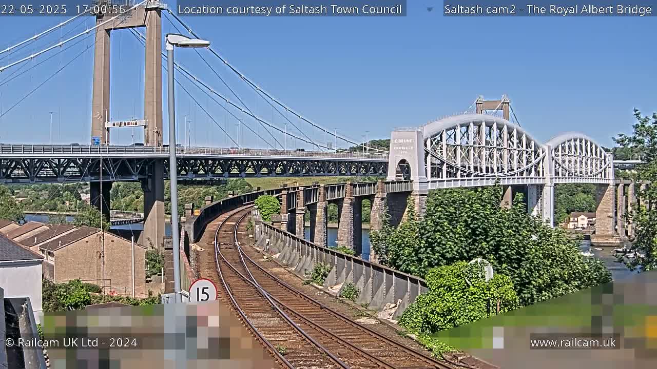 A sunny day view of a railway line running alongside two bridges over a river, one a modern suspension bridge and the other an older, arched bridge.