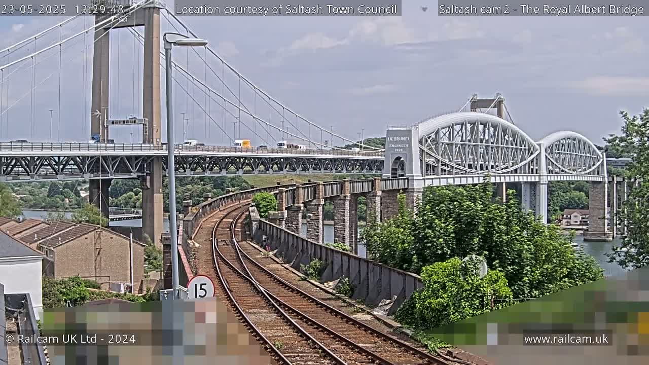 Train tracks curve alongside a river, passing under a large suspension bridge and a smaller arched bridge on a partly sunny day.