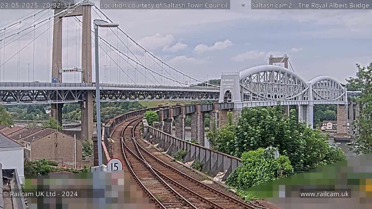 A view of two bridges, one a suspension bridge and the other an arch bridge, over a waterway with train tracks in the foreground under an overcast sky.