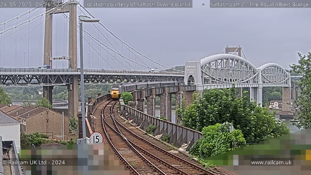 A yellow train travels across a railway bridge under an overcast sky, with another bridge visible in the background.