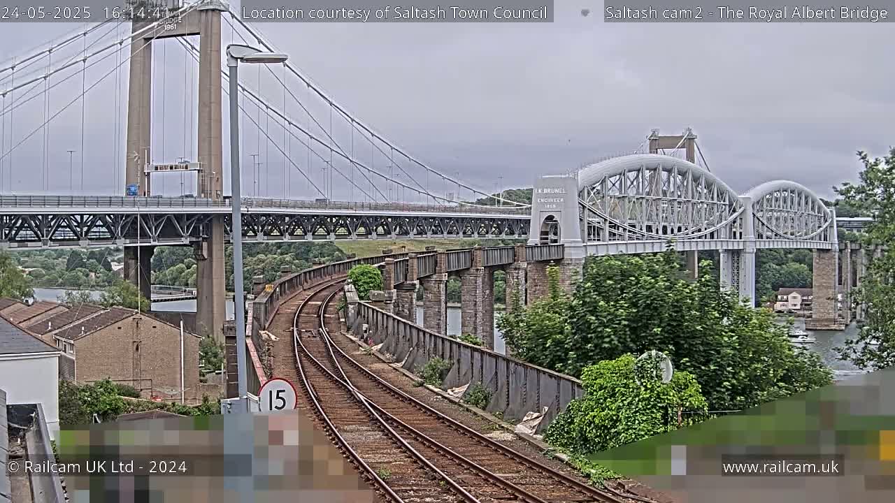 Train tracks curve towards a large suspension bridge and a smaller arched bridge, under an overcast sky.