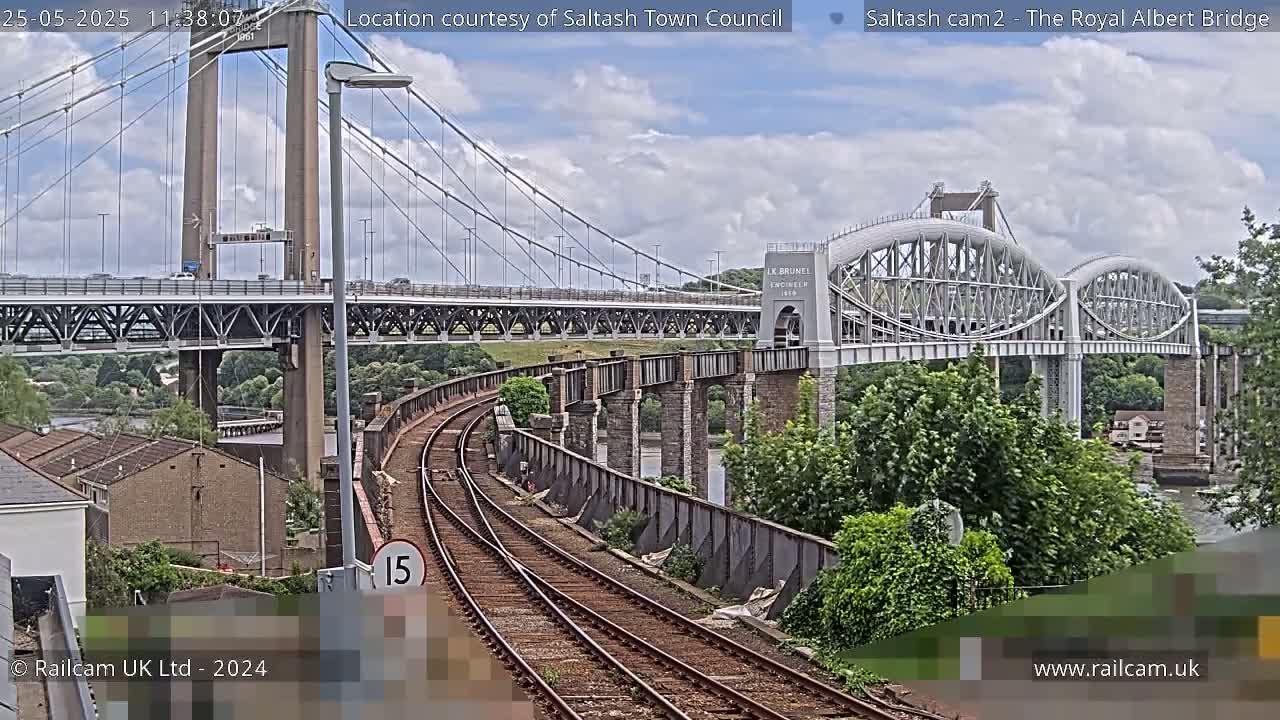 A view of railway tracks leading to two bridges, one a modern suspension bridge and the other an older arch bridge, under a partly cloudy sky.