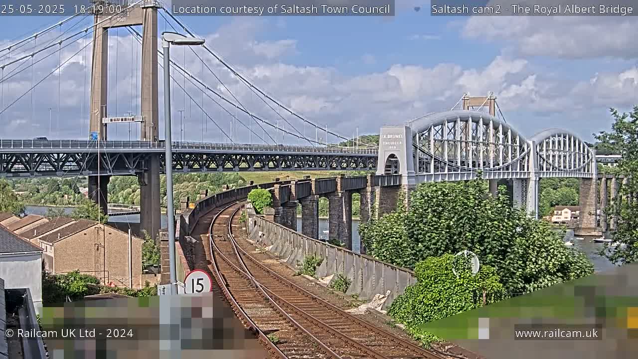 A train track curves alongside a river under a suspension bridge and an arch bridge on a partly cloudy day.