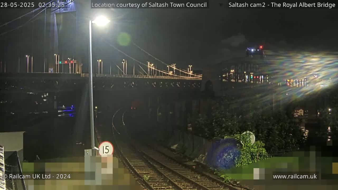 A nighttime view of a suspension bridge and train tracks, with a slightly wet or reflective surface on the tracks.