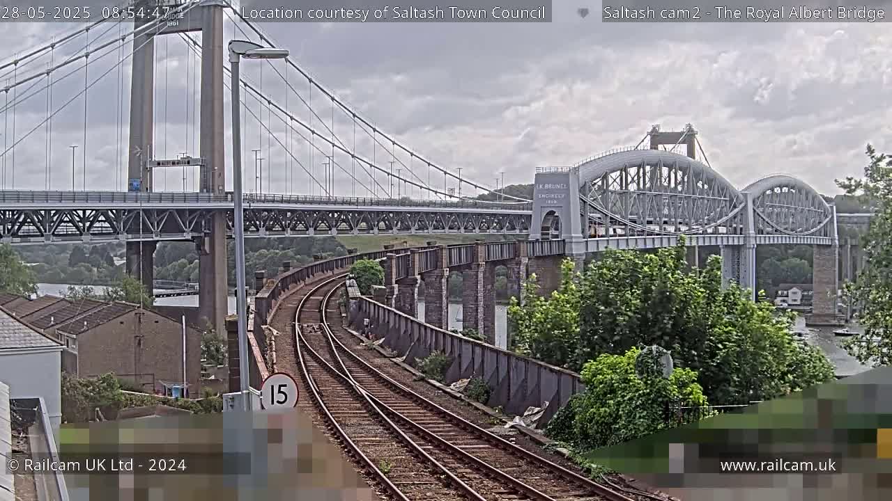 A view of a railway line leading to two bridges, one a suspension bridge and the other an arched bridge, under a cloudy sky.