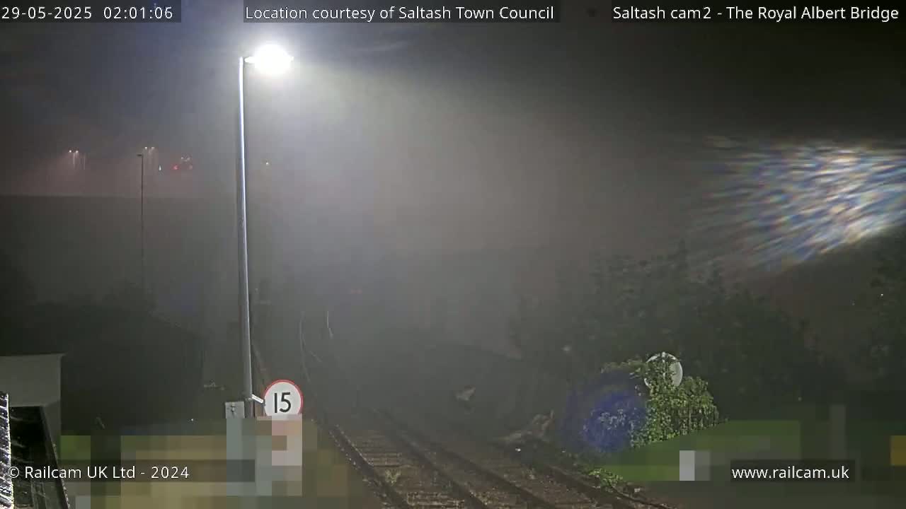 A nighttime scene shows a foggy or misty railway track with a speed limit sign and a tall light post, illuminated by artificial light.