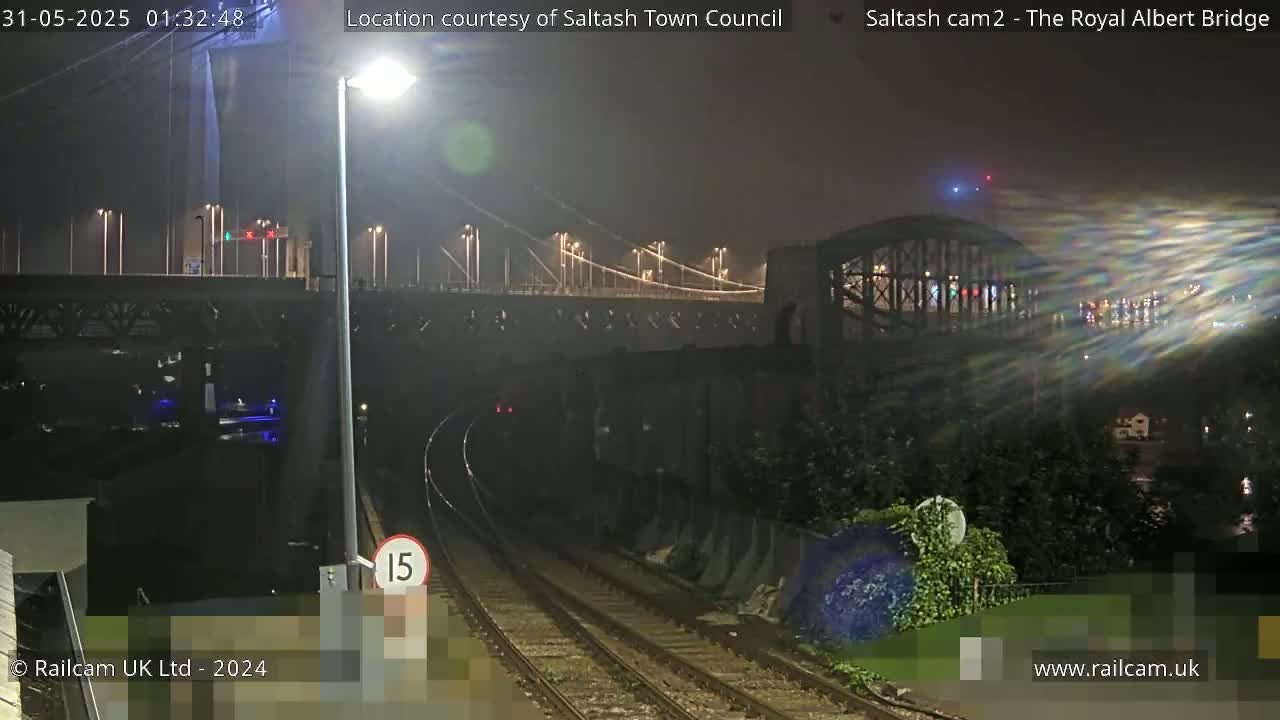 At night, a railway track leads toward a bridge under which a train is passing, with a second bridge visible in the background.