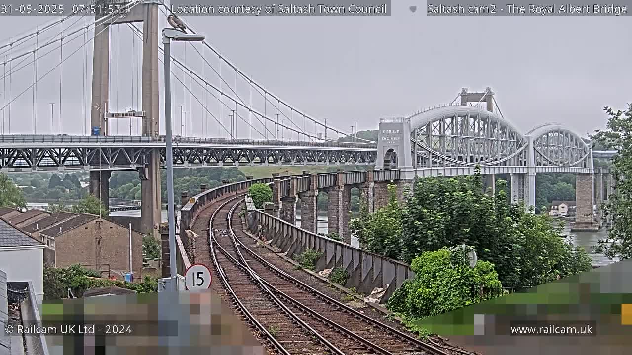 Train tracks curve toward two bridges over water under an overcast sky, one a modern suspension bridge and the other an older, arched bridge.