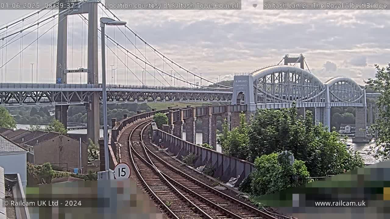 Two bridges, a suspension bridge and an arched bridge, span a river under partly cloudy skies, with train tracks curving towards the viewer in the foreground.