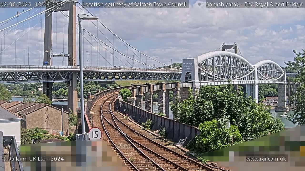 Train tracks curve alongside a river, passing under a suspension bridge and then alongside a large arched bridge on a partly cloudy day.
