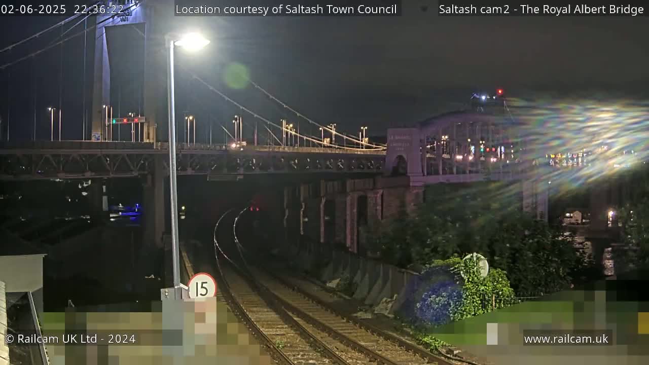 A nighttime view of a train track leading toward two bridges, one a suspension bridge and the other an arch bridge, with vehicle lights visible on both bridges.