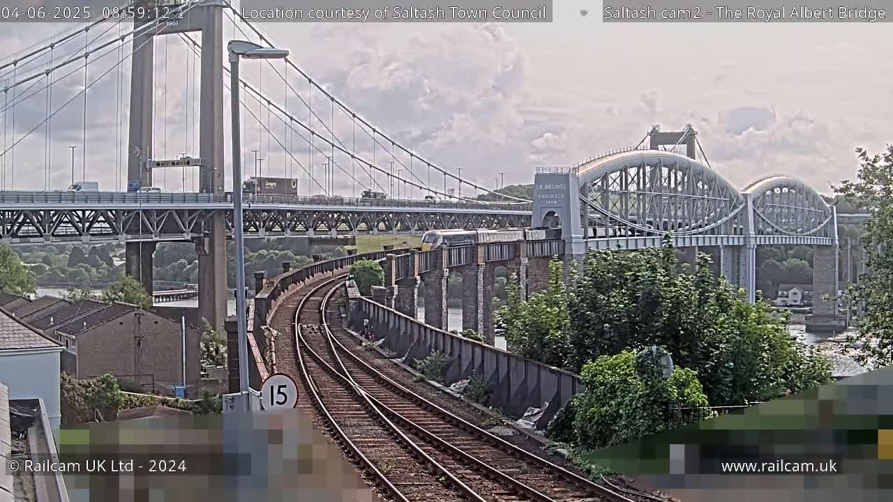 A train passes over a railway bridge, with a larger suspension bridge visible in the background under a partly cloudy sky.