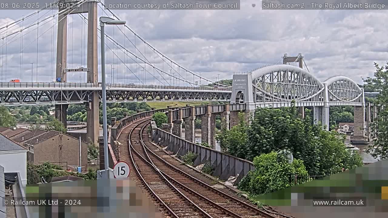 A view of a railway track leading towards two bridges, one a suspension bridge and the other an arch bridge, under a cloudy sky.