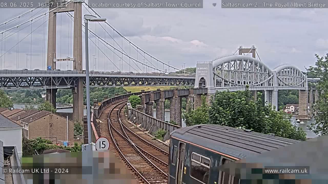 A train approaches a railway bridge, under an overcast sky, with a larger suspension bridge visible in the background.