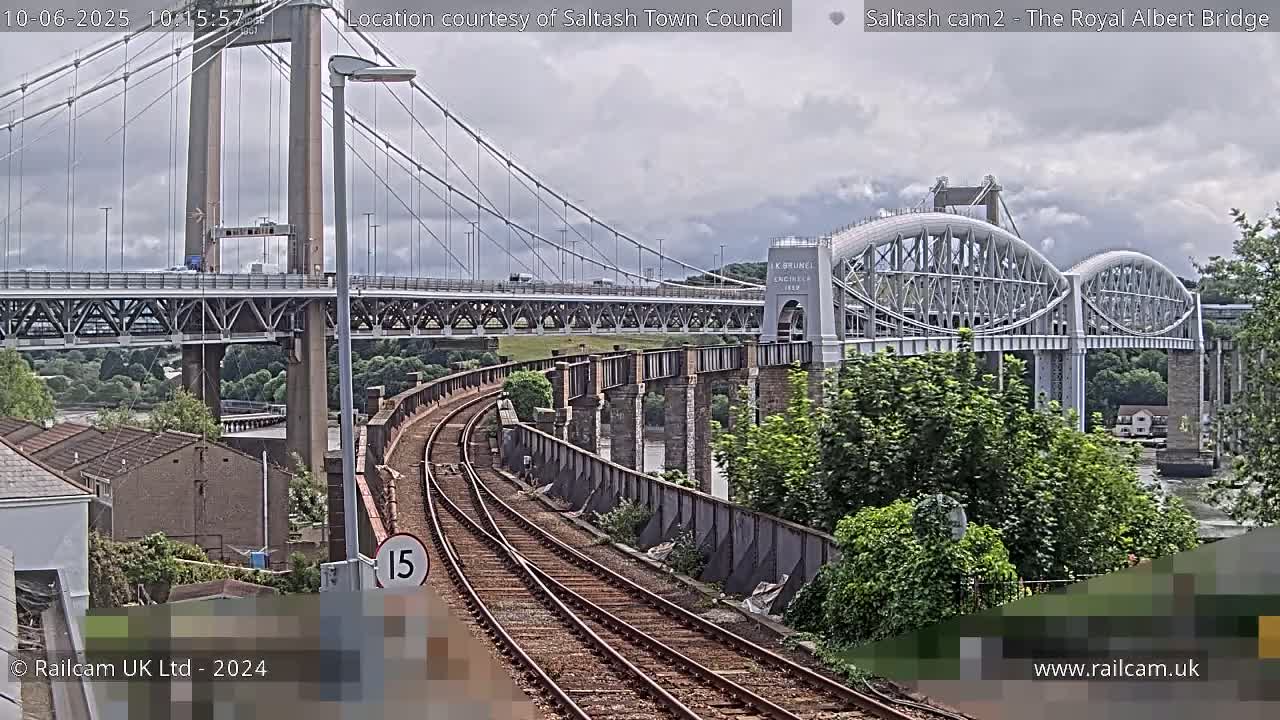 A view of a railway line leading to two bridges, one a suspension bridge and the other an arched bridge, under a cloudy sky.