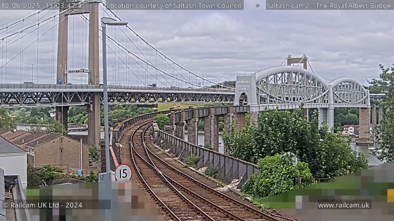 A view of railway tracks leading to the Royal Albert Bridge on an overcast day, with a modern suspension bridge visible in the background.
