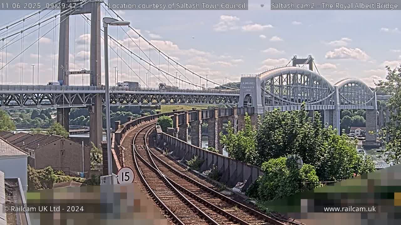 Train tracks curve alongside a river, passing under a suspension bridge and an arched bridge on a partly cloudy day.