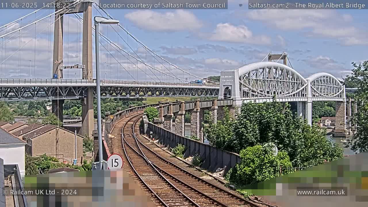 Two bridges, one a modern cable-stayed bridge and the other an older arch bridge, cross a waterway under a partly cloudy sky, with train tracks visible in the foreground.