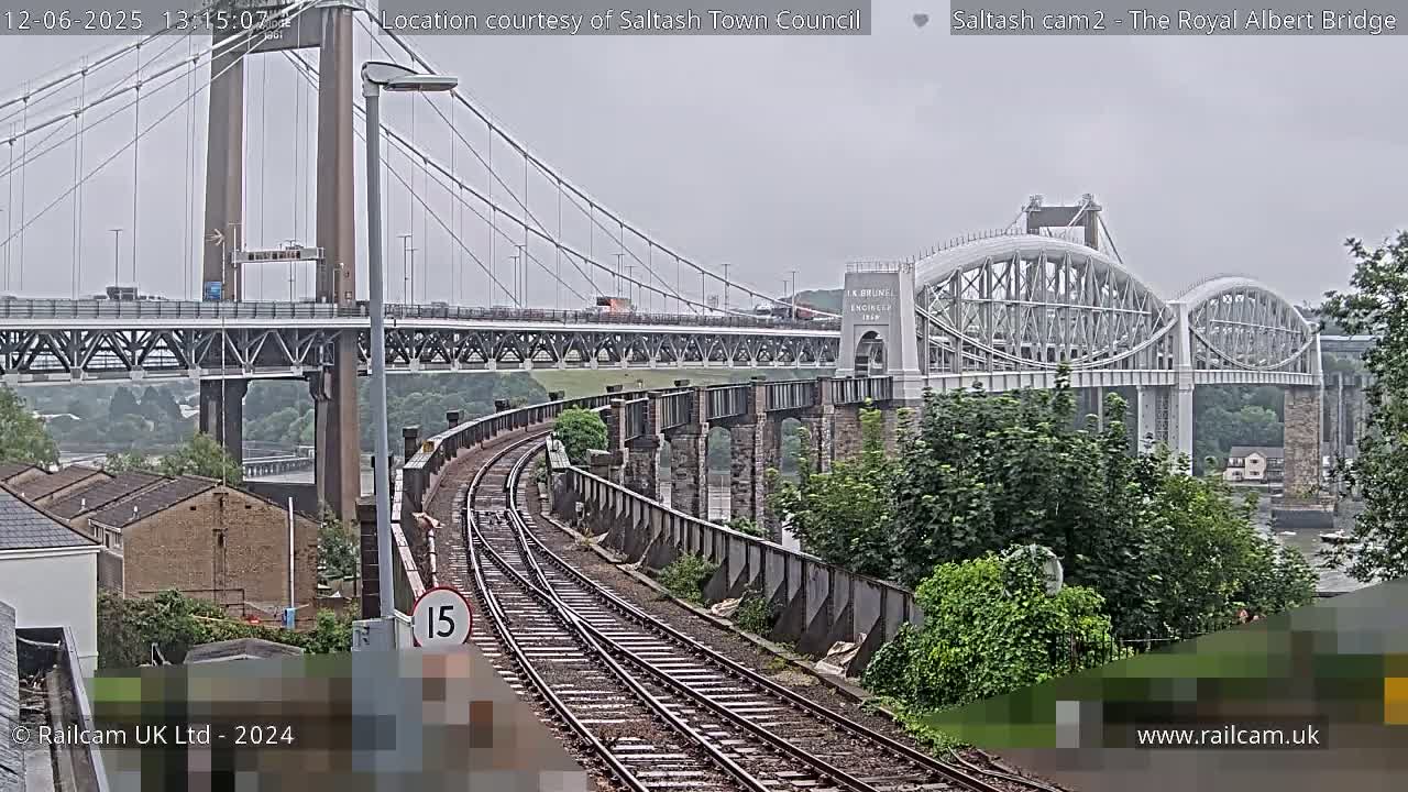 A view of a railway line curving toward two bridges, one a modern suspension bridge and the other an older metal arch bridge, under an overcast sky.