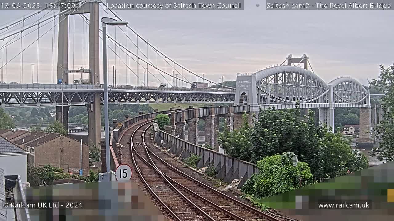 A view of a railway line curving towards two bridges, one a modern suspension bridge and the other a historic arch bridge, on an overcast day.