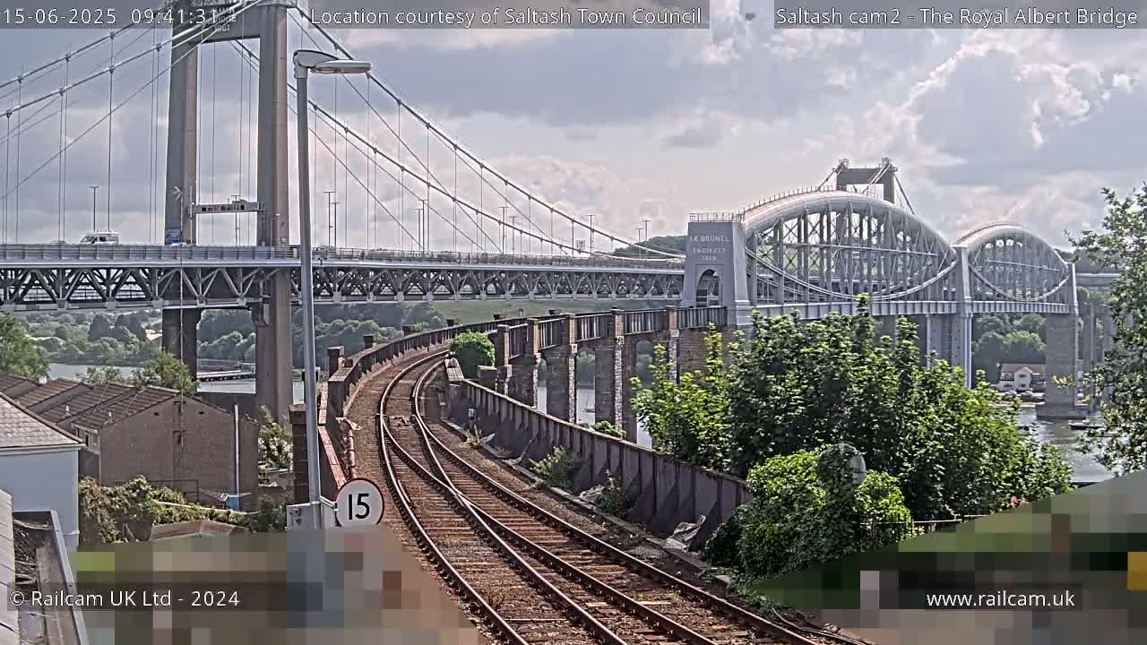 Two bridges cross a river under a partly cloudy sky, with train tracks curving towards the foreground.