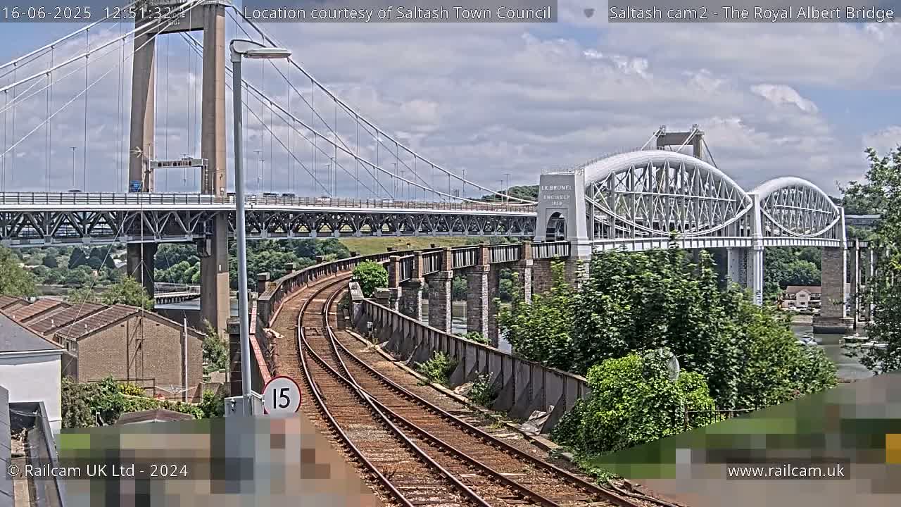 Two bridges, one a suspension bridge and the other an arch bridge, span a waterway under partly cloudy skies, with railway tracks in the foreground.
