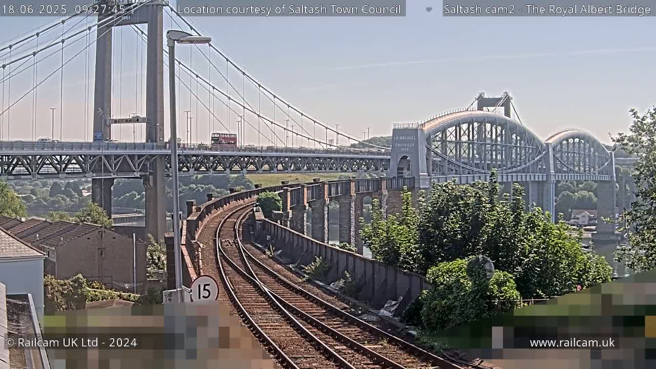 A sunny day reveals a view of two bridges, a modern suspension bridge and an older arch bridge, alongside train tracks curving towards the viewer.