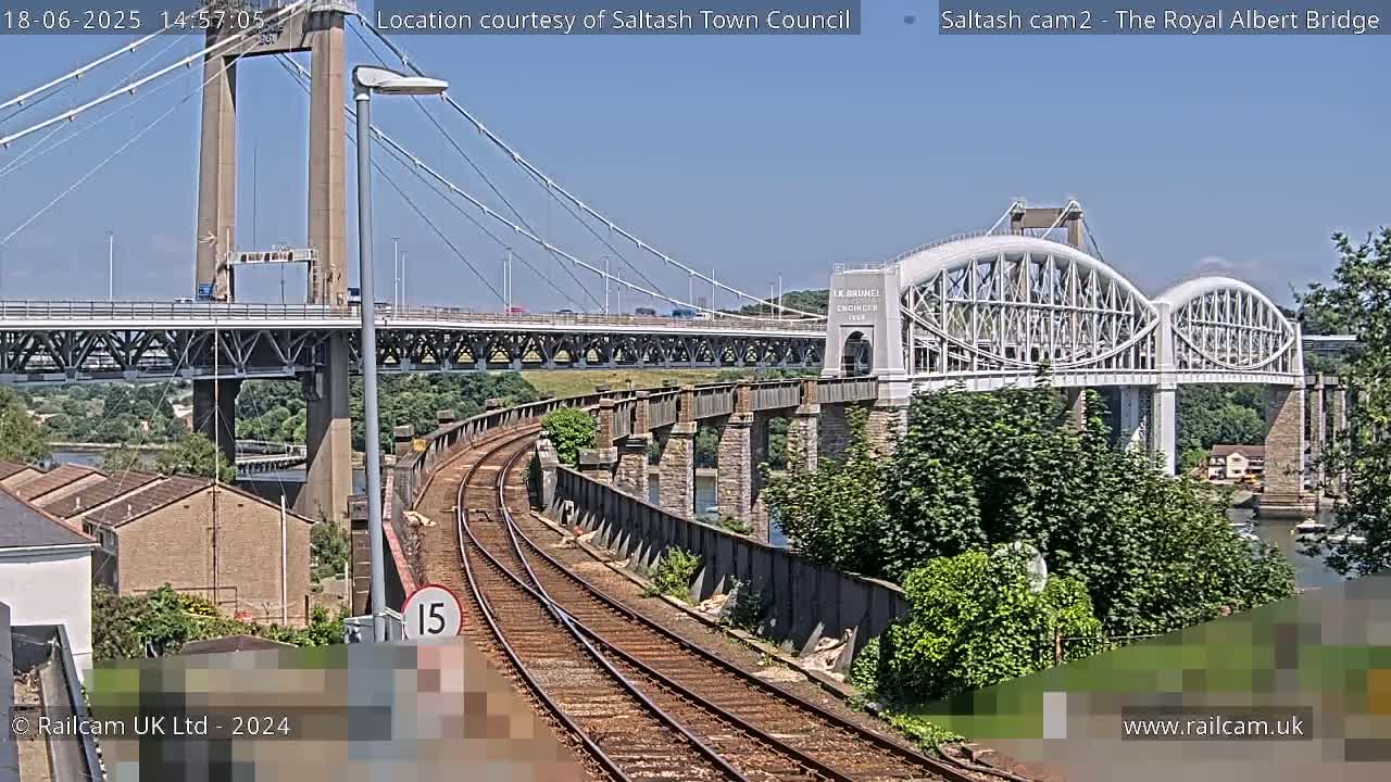 A sunny view of railroad tracks curving towards two bridges, one a modern cable-stayed bridge and the other a historic arch bridge, with lush green vegetation nearby.