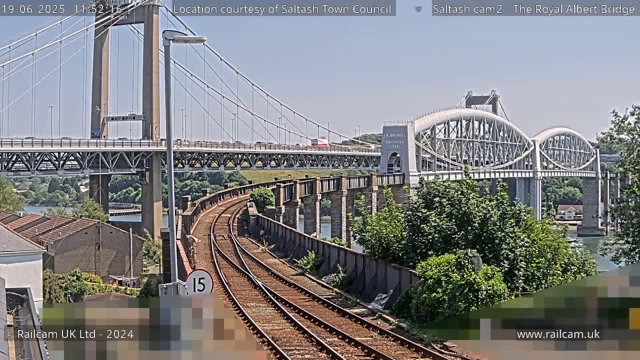 A train track curves alongside a river, passing under a modern suspension bridge and beside an older arched bridge, on a sunny day.