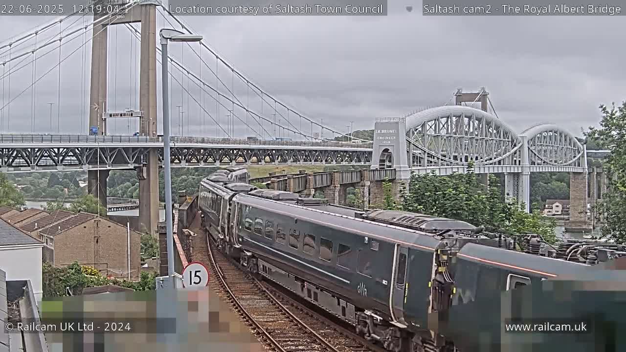A long dark green passenger train travels on a track beside a river, passing under a large suspension bridge and a smaller arched bridge on an overcast day.