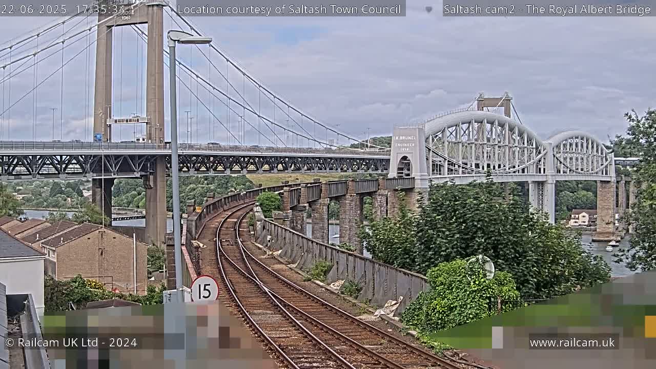 A view of two bridges, one a suspension bridge and the other an arch bridge, over a waterway, with train tracks in the foreground under mostly cloudy skies.