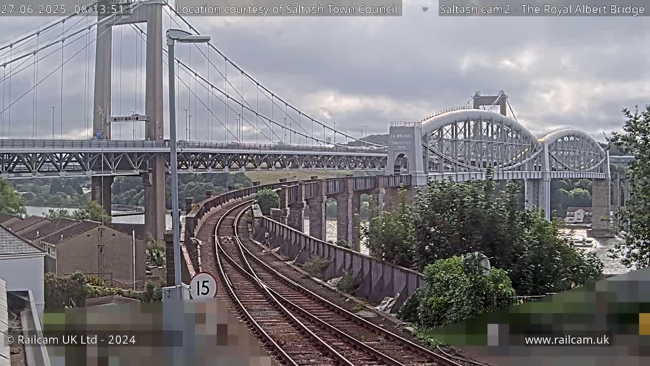 A view of railway tracks curving towards two bridges, one a suspension bridge and the other an arch bridge, under a cloudy sky.