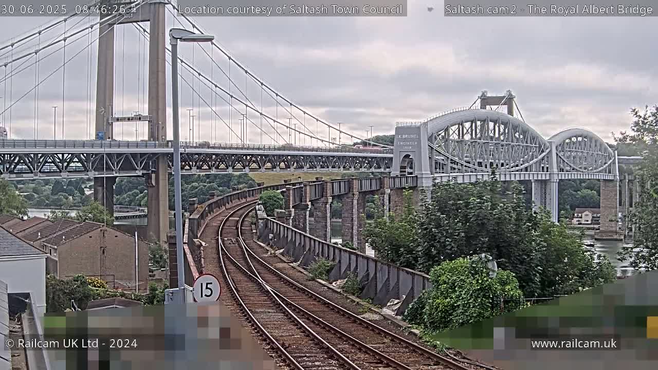 A view of railway tracks leading toward two bridges, one a suspension bridge and the other an arch bridge, under an overcast sky.