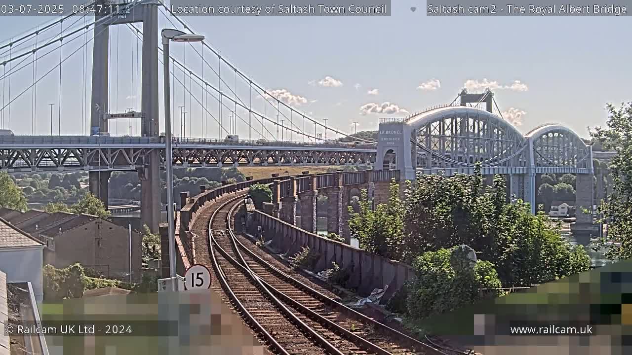 A railway line curves past a section of stone river wall, leading to a view of two bridges on a sunny day.