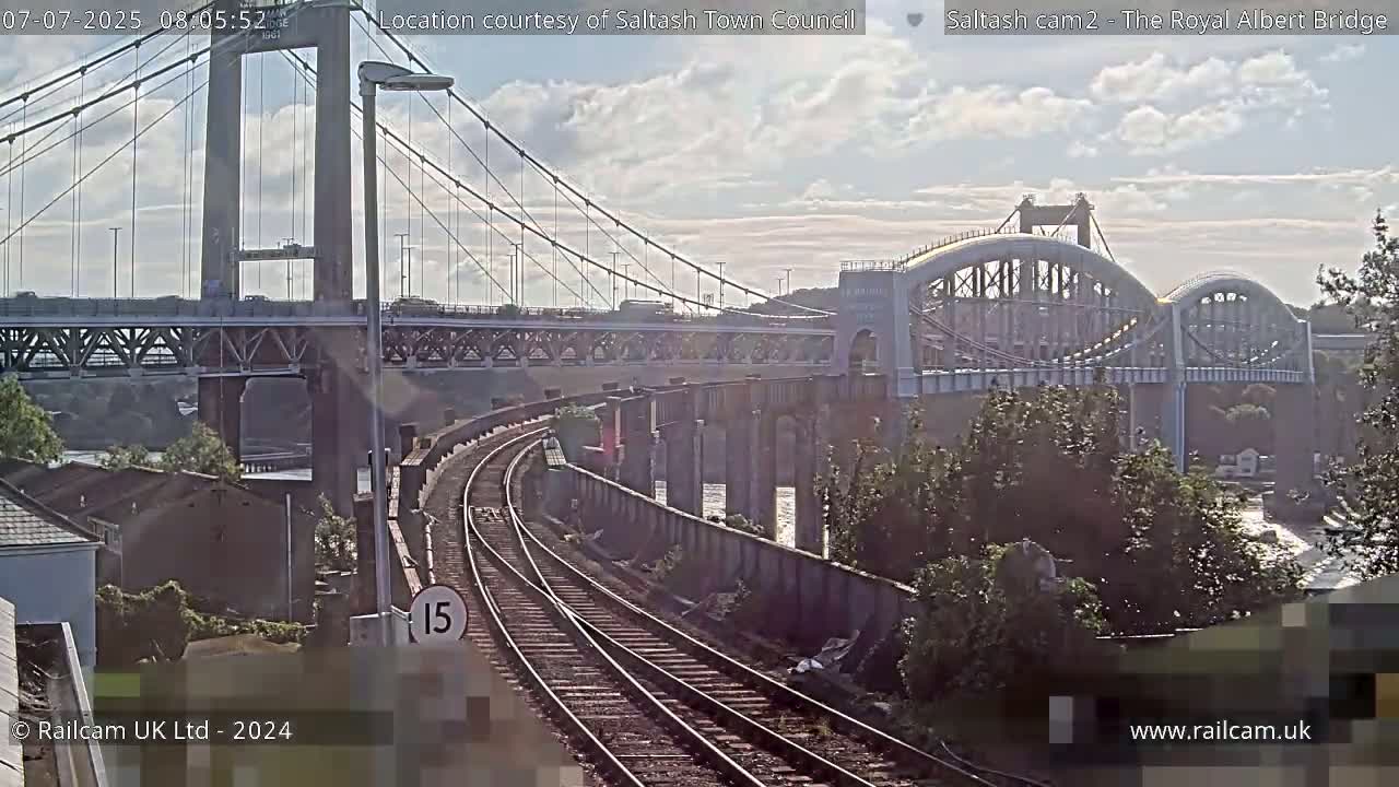 A view of a railway track alongside two bridges spanning a body of water under a partly cloudy sky.